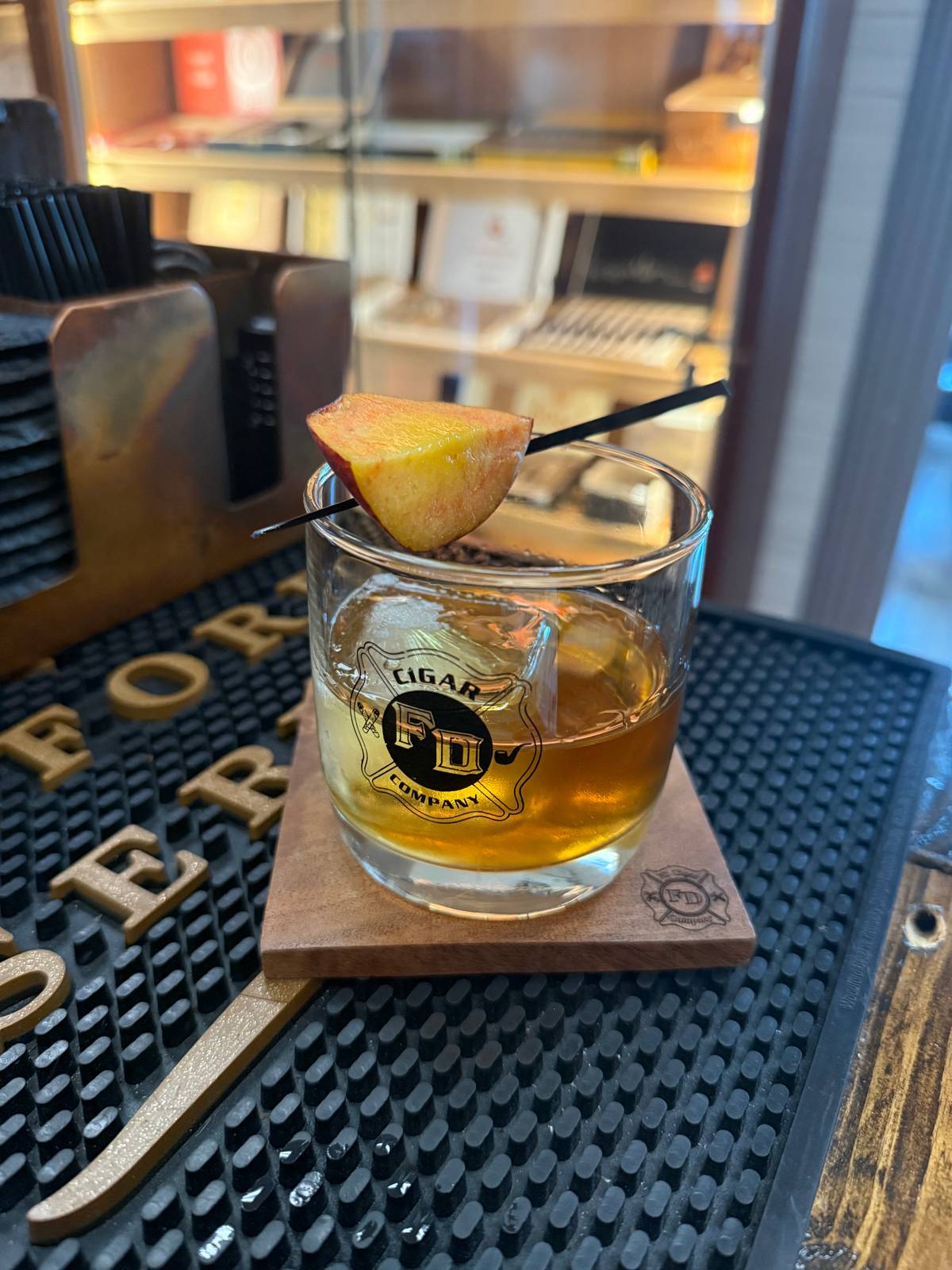 Peach Cobbler Old Fashioned served in an FD Cigar Company rocks glass over a large ice cube, garnished with a fresh peach wedge